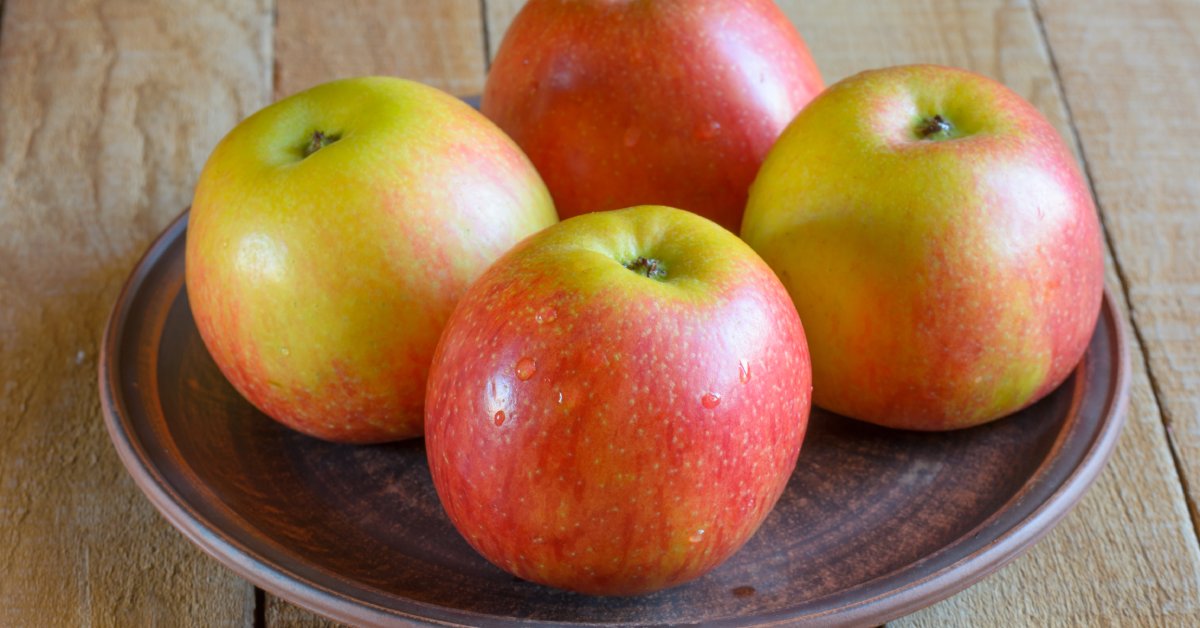 Four large red apples are sitting on a round brown dish. The dish is in the middle of a wooden table.