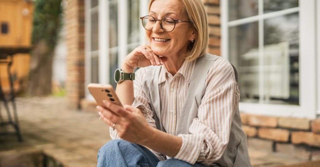 An older woman sitting on her back porch and looking at her phone. She's wearing jeans and a striped shirt.