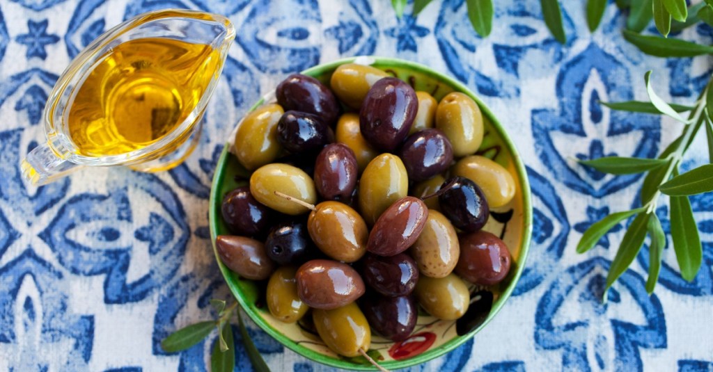 An assortment of fresh olives in a bowl sitting on a blue and white tiled surface. There's olive oil on the left side.