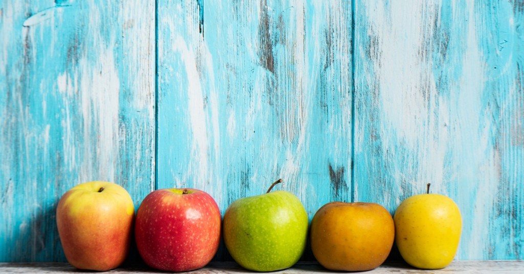Five apples lined up against blue wooden planks. Each apple is a different color, size, and shape.