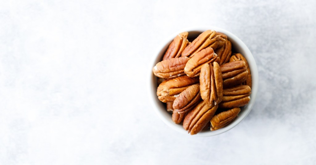 A pile of pecans sitting inside of a small white bowl. The bowl is on top of a white stone countertop.
