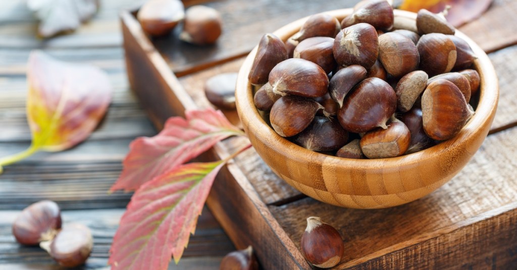 A light brown bowl full of chestnuts. The bowl is sitting on a wooden tray that is on a table outside.