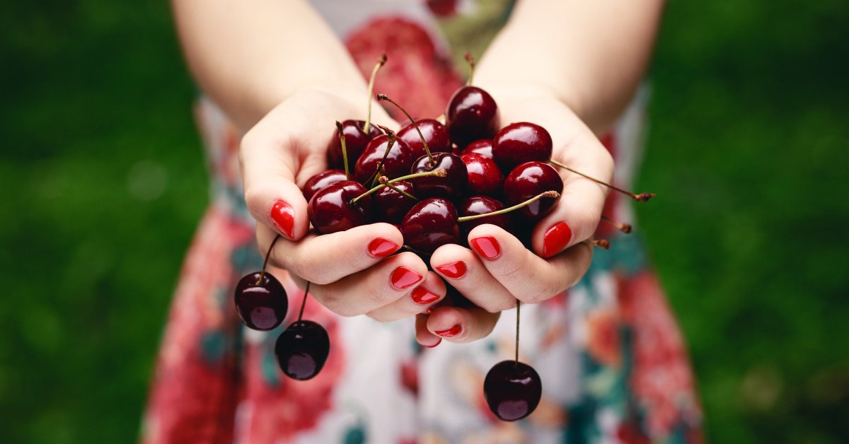 A person with red nail polish wearing a dress and standing outside. They are holding many cherries in their hands.