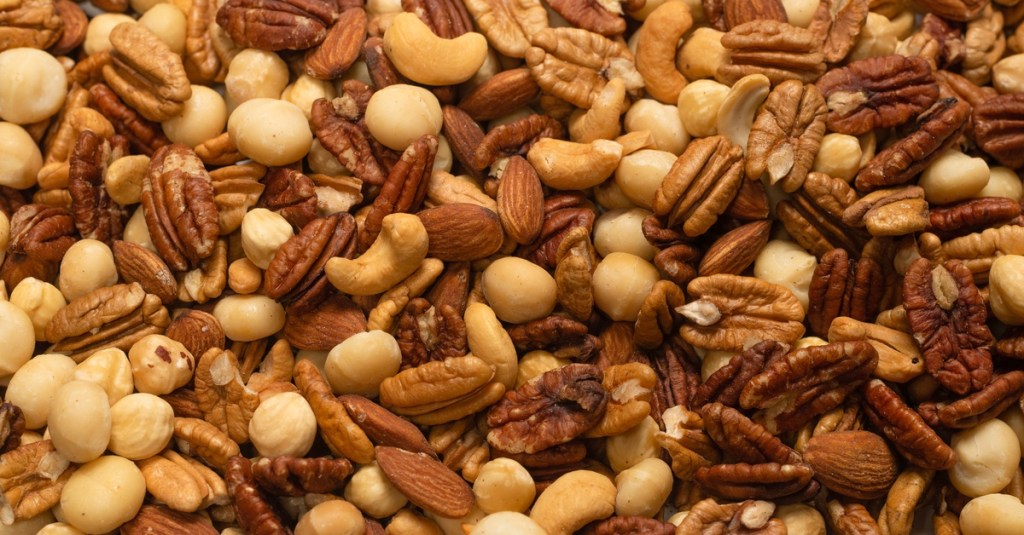 A pile of nuts that includes almonds, pecans, cashews, and hazelnuts. The nuts are in a warmly lit space.