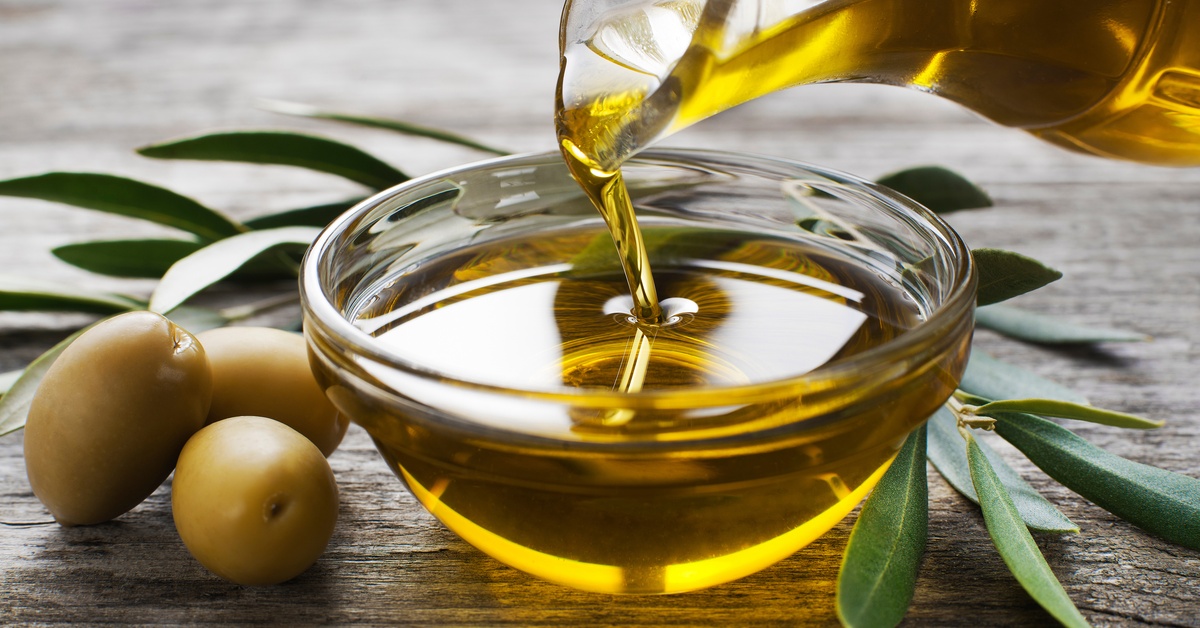 Someone pouring olive oil in a glass bowl on a wooden table. Green leaves and three green olives sit next to the bowl.