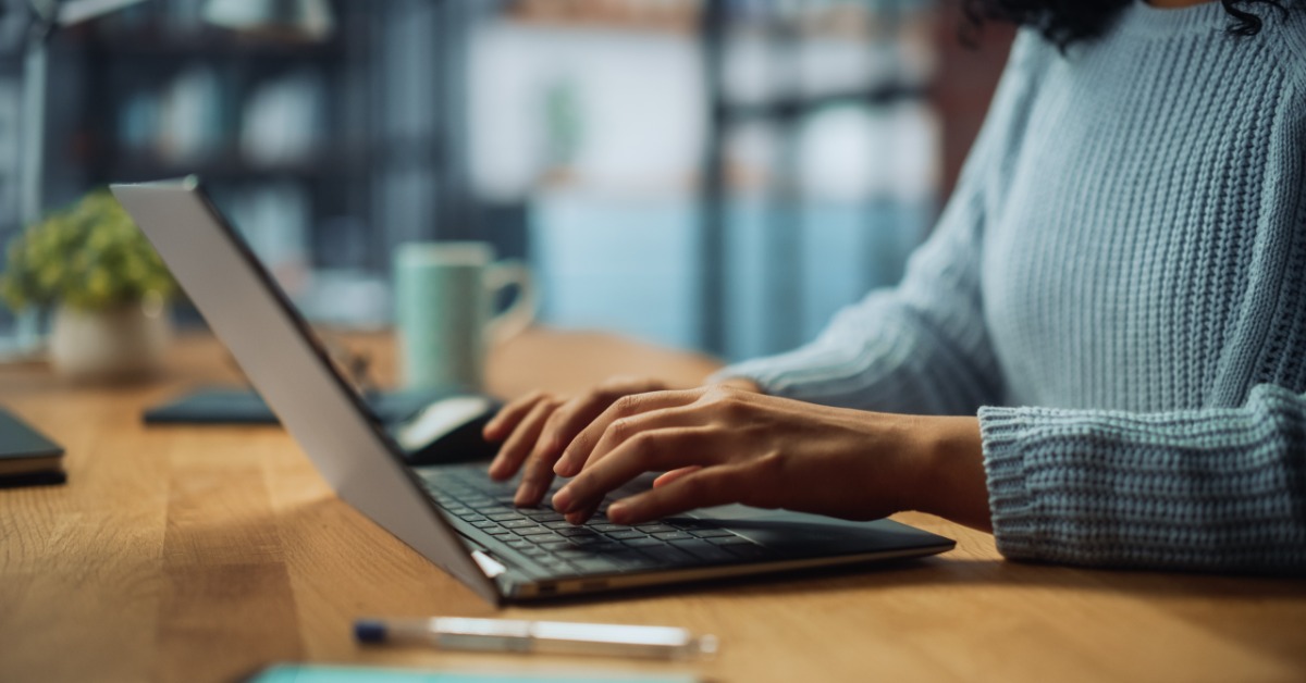 A person wearing a blue sweater tapping on the keyboard of a small black laptop on a long wooden table.