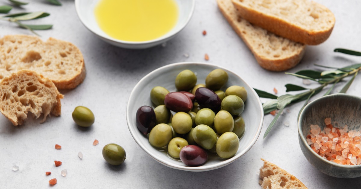 Three bowls surrounded by pieces of bread on a table. One bowl has olive oil, one has olives, and the third has salt.