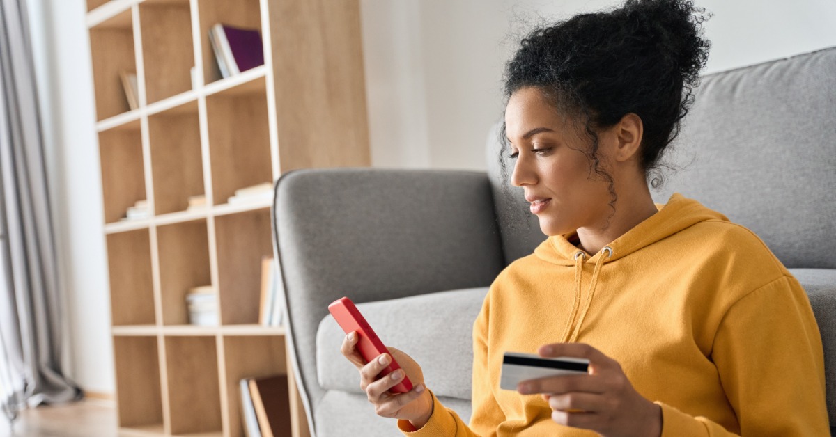 A person wearing a yellow hoodie sitting on the floor holding credit card in one hand and a phone in the other hand.